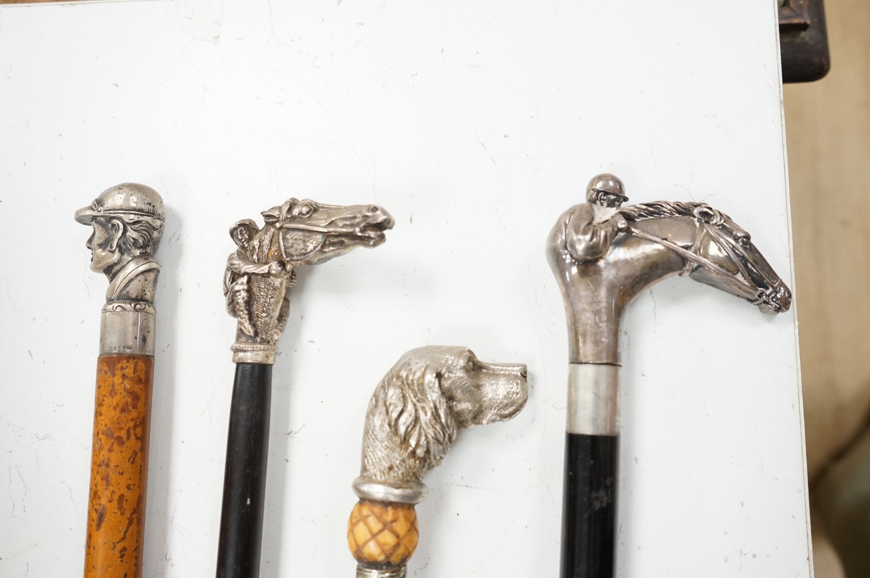 Four 19th century and later walking sticks with silver and white metal tops, to include three horse racing interest examples, largest 96cm in length